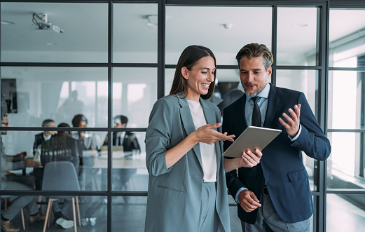 Shot of two coworkers having a discussion in modern office. Businessman and businesswoman in meeting using digital tablet and discussing business strategy. Confident business people working together in the office. Corporate business persons discussing new project and sharing ideas in the workplace.