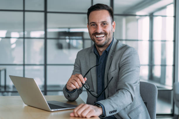Successful businessman in modern office working on laptop. Shot of a businessman sitting on desk in modern office and working on laptop. Portrait of an elegant handsome businessman sitting in his office and using laptop. sales executive stock pictures, royalty-free photos & images