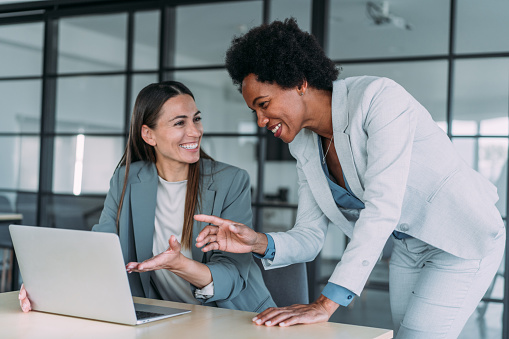 Shot of a two confident business persons sitting on a desk in the office. Two businesswomen in meeting using laptop and discussing business strategy. Business coworkers working together in the office.