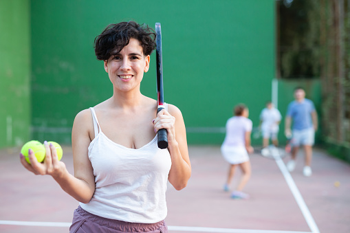 Smiling sporty young Chilean woman ready for friendly frontenis match standing on outdoor fronton court on summer day, holding racket and yellow tennis balls in hands