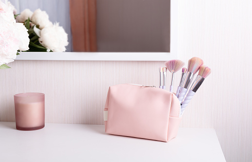 pink cosmetic bag and makeup brushes on a white dressing table against the background of a mirror and a pink decorative candle.