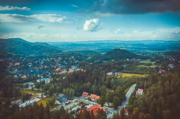 A drone shot of beautiful houses with trees in front of green Karpacz Mountains under cloudy sky