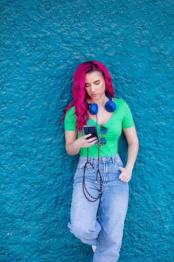 A vertical shot of a beautiful young girl with pink hair and headphones against a blue textured wall