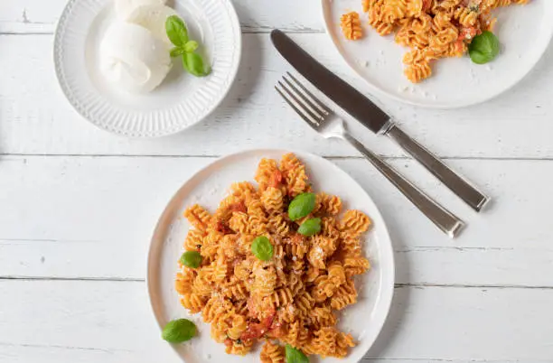 Traditional pasta with fresh tomato, mozzarella cheese. Served with basil and parmesan cheese on a white plate with knife and fork on white table background. Flat lay with space for text.