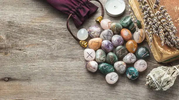 Elder Futhark Rune Stones Set Made of Natural Gemstones with Old Book, Dry Herbs and Sage Incense on Wooden Background with Copy Space