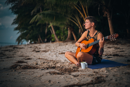 Photo of a young man playing guitar on a beach surrounded by tropical trees