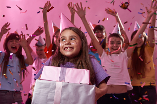 Joyful caucasian little girl with gift boxes with background of celebrating friends in flying confetti. Boys and girls wearing party hats. Holiday and event. Pink background. Studio shoot. Copy space