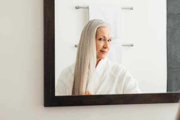 Portrait of an aged woman with long grey hair. Female looking in the mirror admires her hair. stock photo