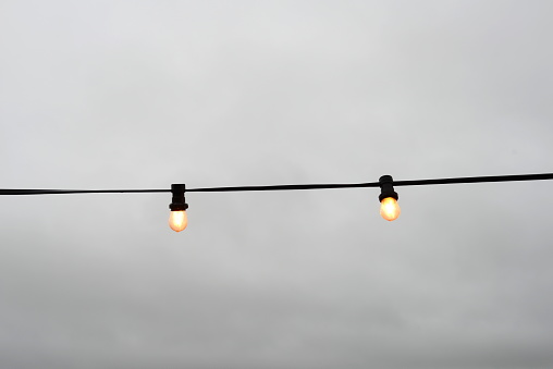 Waving hanging lighting garlands of 2 electric light bulbs on a black electric cable strand in the foreground of a gray rainy sky in winter holidays on the Belgian coast