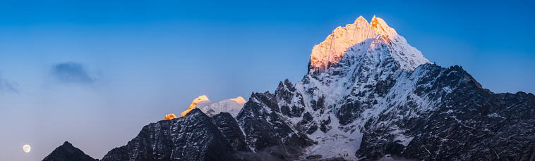 Full moon rising overlooked by the jagged snow capped peaks of Thamserku and Kang Tega spotlit by the golden light of sunset high in the Himalayan mountains of Nepal.