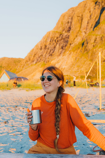 Smiling female with long hair in sunglasses sitting by the table at the sand beach with mountain view during summer night on Lofoten, drinking coffee