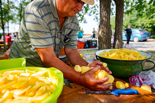 Plastic bowl full of cut young potatoes. Elderly farmer holding in his hands peeled washed raw vegetables over white bowl with water. Outdoor preparing food