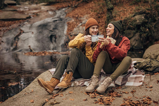 Two women, girlfriends sitting on a rock by the stream together, eating sandwiches and drinking tea.