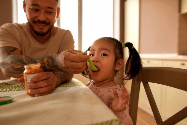 azjatycki ojciec karmi swoją małą córeczkę pokarmem dla niemowląt - sparse healthy eating single father contemporary zdjęcia i obrazy z banku zdjęć