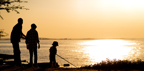 father and sons fishing in the river sunset background. Silhouette of man and two boys fishing at lake. Banner