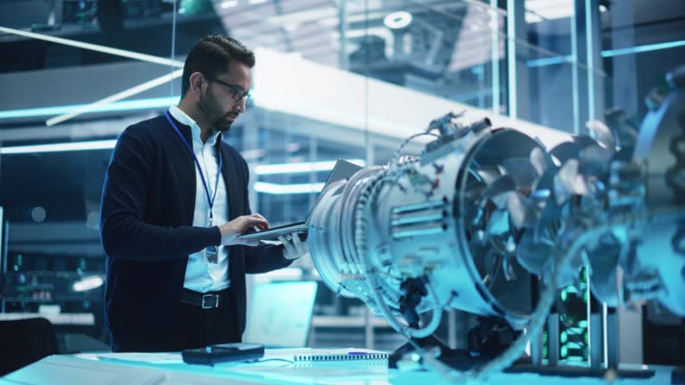 Chief Engineer Using Laptop Computer, Analyzing and Researching How a Futuristic Turbofan Motor Works. Senior Manager Developing Innovative Technology in Industrial High Tech Facility.