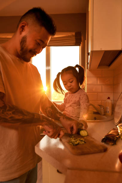 azjatycka dziewczyna biorąc ogórek w pobliżu taty gotującej sałatkę - sparse healthy eating single father contemporary zdjęcia i obrazy z banku zdjęć