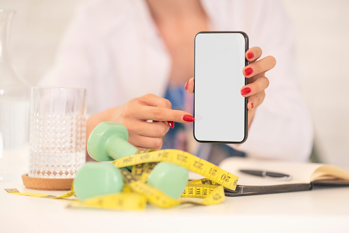 Female dietician with white coat is showing a smart phone which has white screen while she is sitting near her working table. There are two green dumbbells, a glass of water and a tape measure on the white table.