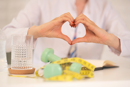 Female dietician with white coat is making a heart shape with her two hands while she is sitting near her working table. There are two green dumbbells, a glass of water and a tape measure on the white table.