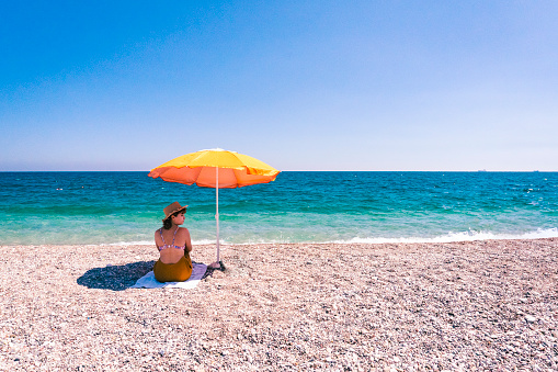 A woman in a fedora enjoys a vacation under an orange parasol at the beach on a summer day.