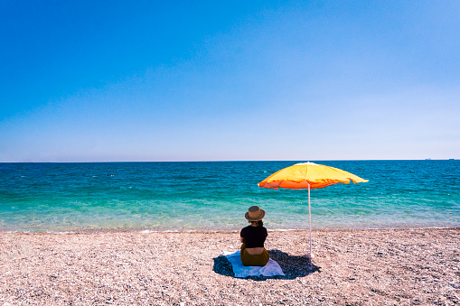 A woman in a fedora enjoys a vacation under an orange beach umbrella at the beach on a summer day.