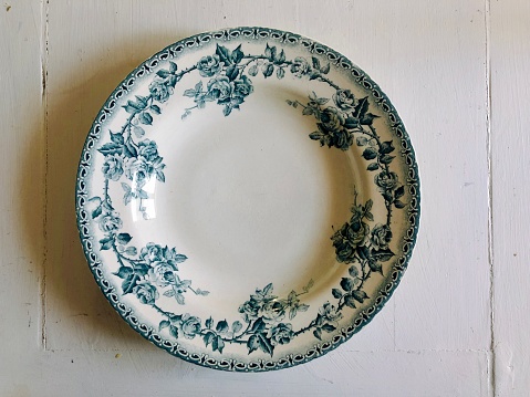 Horizontal high angle closeup photo of a round white porcelain plate with a blue leaf and rose flower pattern around the edge, on a rustic white painted wooden table in a dining room. Camargue, Provence, south of France.