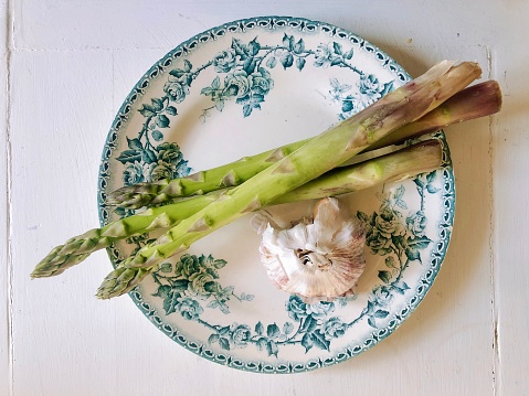 Horizontal high angle closeup photo of three freshly harvested asparagus spears and a bulb of garlic on a white ceramic plate with a blue leaf and rose flower pattern around the edge, on a rustic white wooden table. Camargue, Provence, south of France.