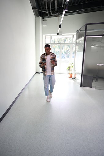 Full-length of African American young man using mobile phone while walking along office hall, holding cup of drink