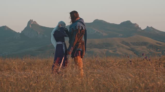 A couple in love, a man and a woman in medieval costumes, against the backdrop of a mountain landscape in the rays of a sunset in the mountains. Slow motion Side view