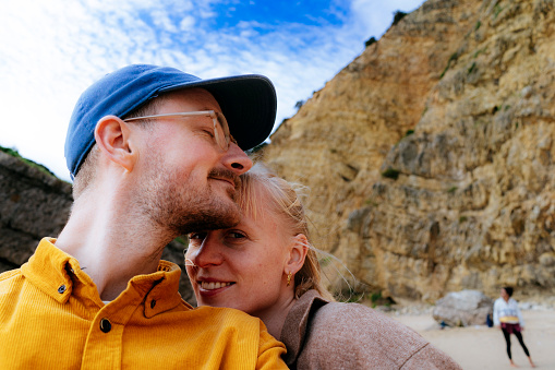 A happy millennial couple takes a selfie on a beautiful sandy beach in Portugal. They are on vacation, enjoying the warm sunshine and crystal clear water. The image captures the young couple's carefree and relaxed spirit, making it perfect for use in travel and vacation-related content.