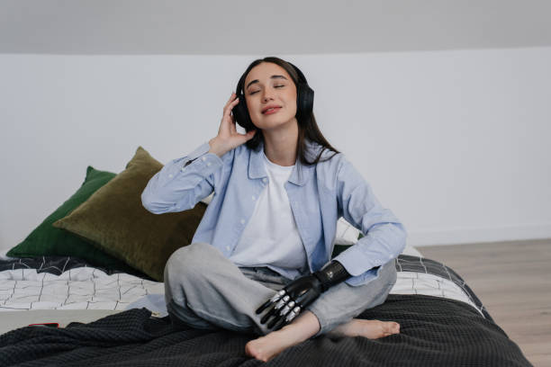 istock Young woman sitting on bed at home, with  bionic hand prosthesis, smiling, relaxing after remote learning. Attractive hispanic girl in headphones starting day, listening music at bedroom. Orthopaedics 1454610635