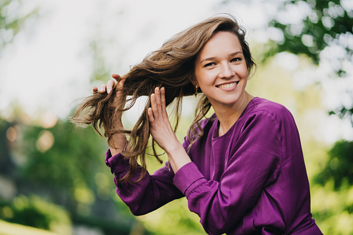 Beautiful young woman with long hair dressed in purple dress holds her hair, walking outdoors, broad toothy smiling. Happy people concept. Close up of gorgeous woman on blurry nature background.