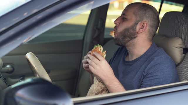 Man driver eating a burger at the wheel of a car. Unhealthy fast food. Concept of street food. Food in travel