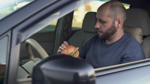 Serious man eating fast food while driving a car. The taxi driver eating a big juicy burger