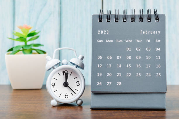 The February 2023 Monthly desk calendar for 2023 year with alarm clock on blue wooden background. stock photo