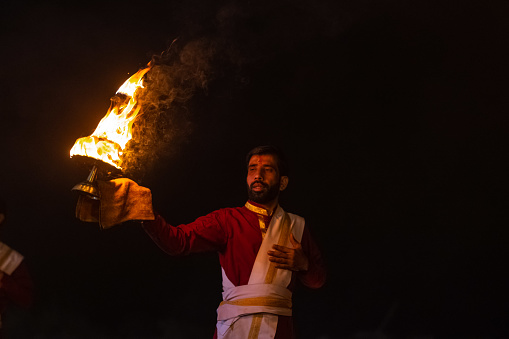 Rishikesh, Uttarakhand, India - October 2022: Portrait of hindu male priest performing river Gange aarti with fire flame in hand at triveni ghat to worship river ganges in rishikesh. Ganga aarti performed in night near river ganges bank.