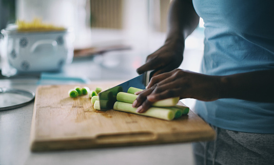 Unrecognizable woman cutting leek on the wooden cutting board in her kitchen at home, preparing an organic vegetarian meal.
