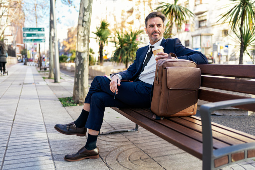 Handsome middle aged male entrepreneur in classy suit sitting on bench with coffee to go in city and looking away