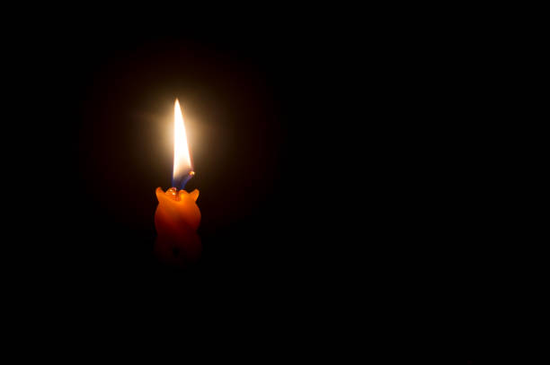A single burning candle flame or light is glowing on an orange candle on black or dark background on table in church for Christmas, funeral or memorial service with copy space.