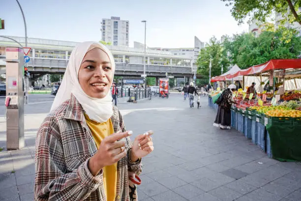 young smiling muslim woman with hijab standing on street in berlin kreuzberg in front of street market