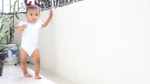 beautiful baby latina with brown skin, taking her first steps. curious little girl walking on the balcony, holding on to the wall learning to walk.
