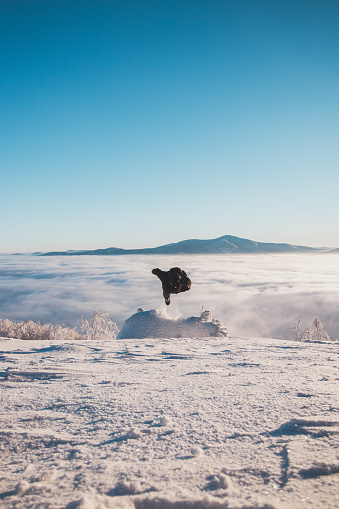 Energetic boy in winter clothes jumping from a stump into the fresh snow with a view of the sunrise and clouds rolling along the mountains. Beskydy mountains, Czech Republic.