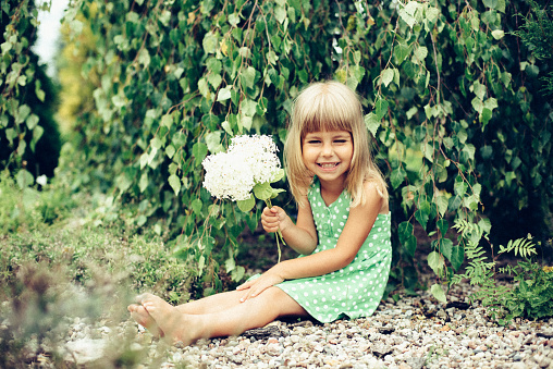 A little girl in a beautiful dress with flowers and short blonde hair enjoys beautiful flowers in her grandmother's garden in the village