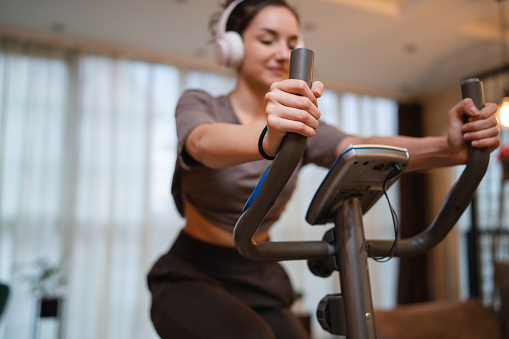 One woman young caucasian female training at home with headphones on Indoor Cycling stationary Exercise Bike real people copy space health and fitness workout concept selective focus on hand