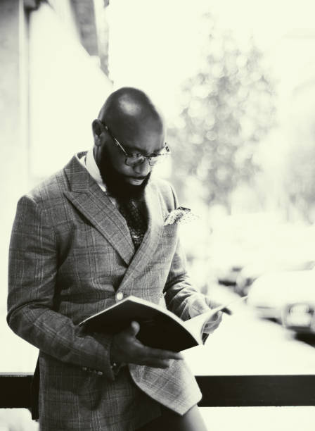 A vertical black-and-white portrait of a dapper bearded African American businessman reading a magazine outdoors; a greyscale shot of an elegant black guy in eyeglasses and a tailored checkered suit