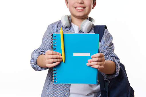 Focus on a blue copybook with yellow pen in hands of a cheerful teenage school boy, wearing casual wear, smiling a toothy smile, isolated on white background, copy space. School and education concept