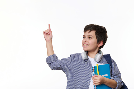 Cheerful dark-haired multi-ethnic teenage boy in casual wear, pointing at copy space for advertising on white background, posing with copybook and school backpack. Back to school. Knowledge concept