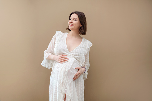 Attractive pregnant woman in white dress on beige background. Motherhood and healthcare concept