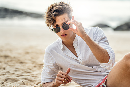 Portrait of handsome young italian man relaxing at the beach, wearing white shirt and fashionable sunglasses. Summer time.