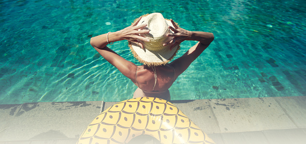 Back view of young woman wearing summer hat relaxing in big swimming pool with blue water on a sunny day. Copy space. Luxury hotel.